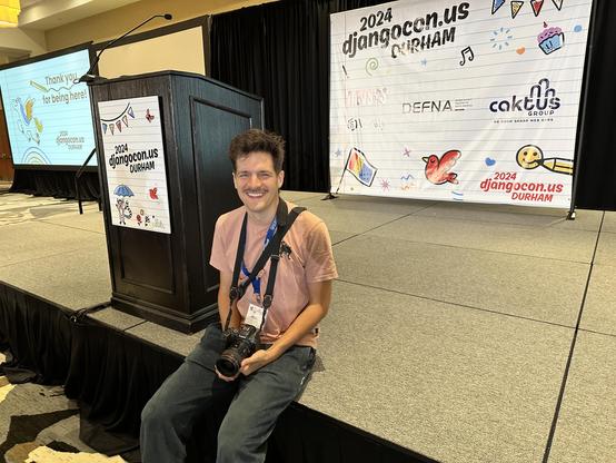 DjangoCon US photographer Bartek Pawlik sitting on stage in front of the backdrop.