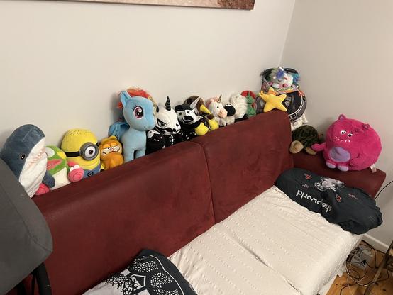 The back rest of a red couch stacked with all sorts of plushies. Unicorns, blahaj, Garfield, minions and sheep to name only a few.
