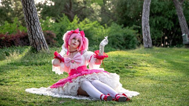 A cosplay of Madoka Kaname from the anime Puella Magi Madoka Magica in the dress that appears on the Concept Movie Trailer. Madoka sits gracefully on the grass with a calm and seren expression. The lush greenery and soft sunlight in the background give the scene a peaceful and magical atmosphere, capturing the essence of the character.