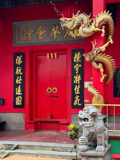 Rote Tür an chinesischem Tempel in Kuala Lumpur. Mit sich um eine rote Säule windenden, goldenem Drachen, Steinhundedrachen und chinesischen Zeichen um die Tür herum. Die türknäufe und der Verschlussriegel bilden ein genervt blickendes Gesicht