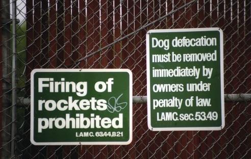 Two green signs on a fence. The left sign states "Firing of rockets prohibited," while the right sign reads "Dog defecation must be removed immediately by owners under penalty of law."