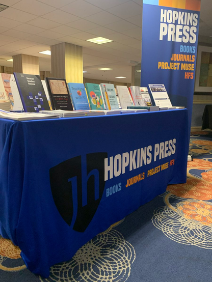 Photo of the Hopkins Press table at German Studies Association 2024 conference. A blue tablecloth with the Hopkins Press logo draped over a table, topped with a display of books and journals from the Hopkins Press collection