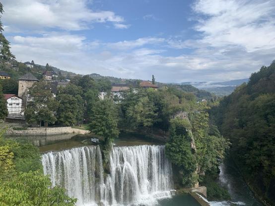 A forested valley with a town beyond the trees, and a broad, high waterfall under a party cloudy sky