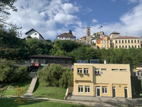 A steep ridge with buildings at the bottom, middle, and top, and a fortress with tower and Bosnia-Herzegovina flag
