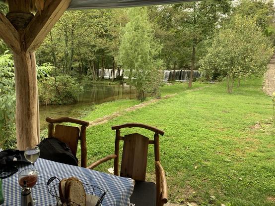 A table with wooden chairs and blue gingham tablecloth. In the background, trees, a river, and a broad, low waterfall