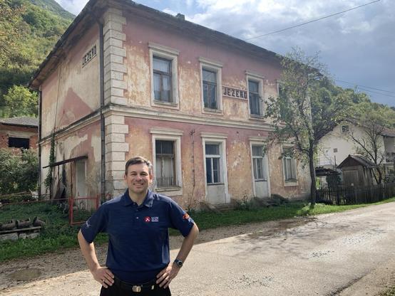 A man in a shirt with the words “Mars 2020 Perseverance” stands in front of a two-storey house  with signs on it reading “JEZERO”