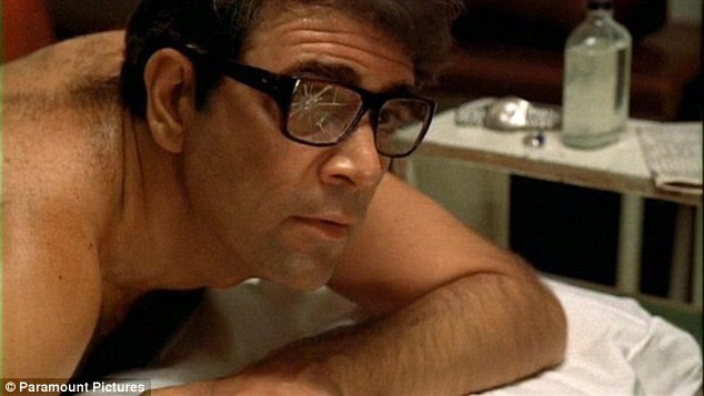 Man lying shirtless on a massage table, wearing dark-rimmed glasses. The right lens of the glasses is cracked. His facial expression is neutral, with eyes looking off to the side. In the background, a glass bottle, a watch, and a newspaper rest on a table. This image is a still from the movie "The Godfather," showing the character Moe Greene, portrayed by actor Alex Rocco.  [Alt text by ALT Text Artist GPT]