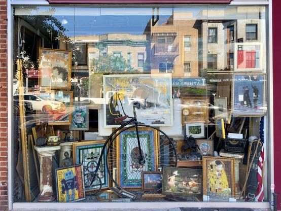 Cluttered but pleasing window display for a framing shop containing paintings, posters, and a toy unicycle. The buildings across the street appear in the reflection.
