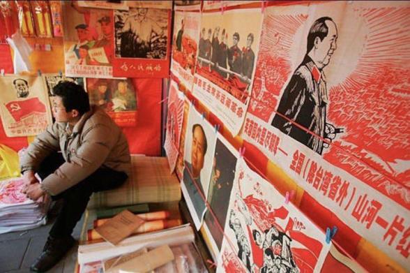 A man sitting in a shop with dozens of Mao Zedong posters on the walls.