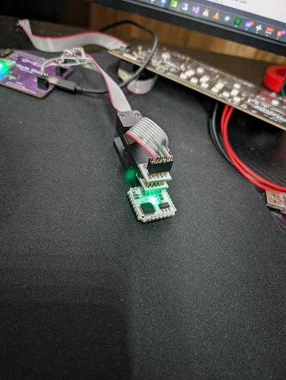 A small circuit board with a glowing led is connected to a pogo-pin debug interface.
Other boards and a monitor can be seen in the background