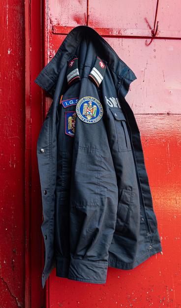 A black firefighter’s jacket with various badges and patches sewn on it, hanging on a hook against a red painted door.