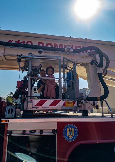 A young girl in a pi k dress and wearing a firefighter’s helmet standing on the platform of a cherry picker on a white and red fire engine. The Sun is shining in a blue sky behind.