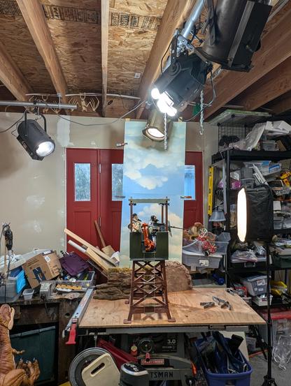 Workshop/studio with a three foot tall miniature guard tower sitting against a sky background with fake rocks at the base. Theatrical lights on a suspended ceiling grid and on a stand illuminate the scene. Three characters, represented by costumed 12" action figures and a 1/6th scale robot arm, occupy the cupola at the top of the tower. 