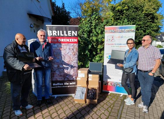 In einem Vorgarten stehen 4 Personen, von links nach rechts Donaly Malungu, Flugpate und Labdoo-Helfer, Ralf Hamm, Labdoo.org und Michaela und Michael Roos von Brillen-ohne-Grenzen.
Dazwischen Kartons mit gespendeten Brillen und Sonnenbrillen sowie je ein Rollup von Brillen ohne Grenzen und Labdoo.
Der Himmel ist strahlend blau, im Hintergrund Bäume und Büsche. 