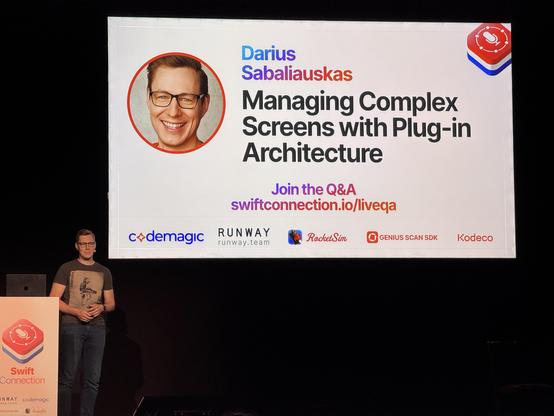 Man standing on stage during presentation. Presentation says "Managing Complex Screens with Plug-in Architecture"