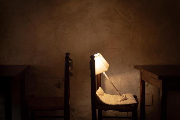 Fotografía donde se ve un interior iluminado tenuemente por una lámpara que está apoyada en una silla de tal manera que parece que está ahí sentada descansando.