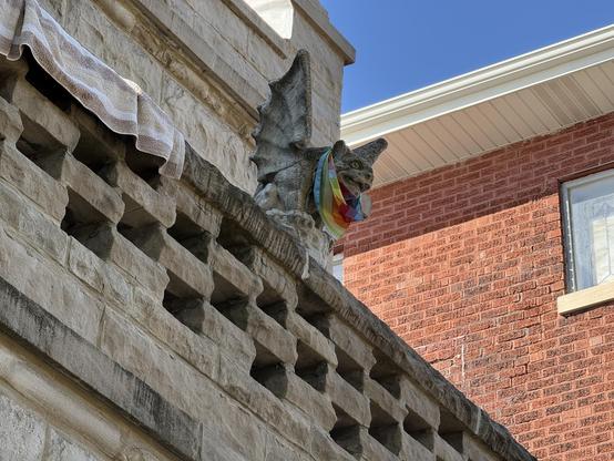 A stone gargoyle wearing a rainbow scarf is perched on the edge of a building, with a towel draped over the ledge above it. In the background, a red brick wall and a window are visible under a clear blue sky.