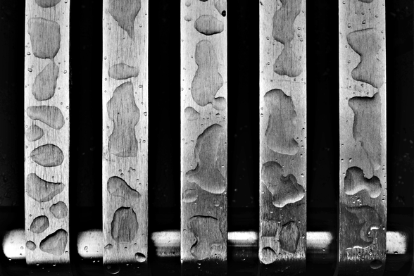 A high contrast photo of a stanless steel bench. The silver veertical slatts of the bench are dotted with large rain droplets. The gap inbetween is pure black. The structure of the metal can been seen in detail whilst the water puddles magnidy the metal structure in parts.