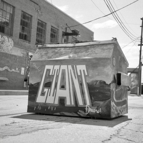 A black and white film photo. A large dumpster has a hand-painted message that states “Have GIANT Dreams!” in Paintbrush Alley in San Angelo, Texas. Sunday, September 1, 2024.