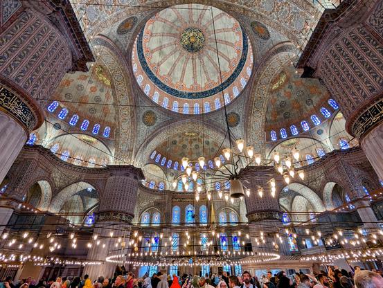 A breathtaking upward view of the Blue Mosque's interior in Istanbul. The image showcases the mosque's magnificent central dome adorned with intricate designs and calligraphy. Sunlight streams through stained-glass windows, illuminating the vast prayer hall filled with worshippers.