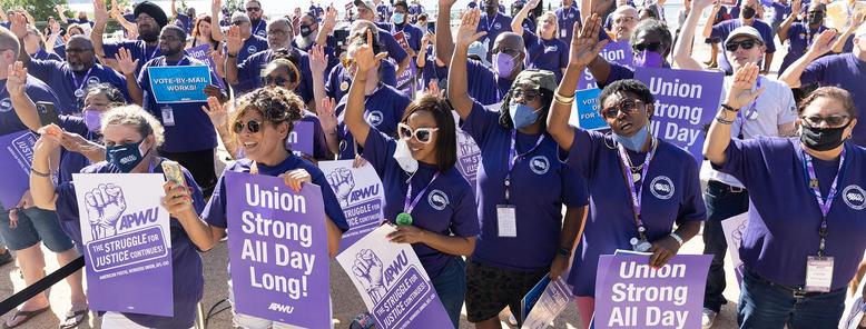 Postal workers union members rally holding Union Strong All Day long signs and APWU purple shirts