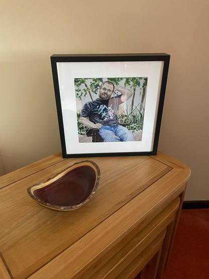 ID Framed print of a portrait of Scott who is wearing a tshirt and jeans an sat in a wheelchair in an outdoor environment. The print is standing in on the corner of a pine table with a wooden bowl in front of it