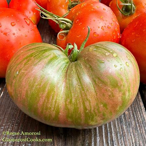 This photo shows a Metallica Tomato, a pink and green striped variety from the Dwarf Tomato Project, an initiative described on their website as an “all volunteer world-wide tomato breeding project.” The Metallica tomato is on a weathered wood table with red Big Boy tomatoes in the background. Photo ©Reese Amorosi for GlamorosiCooks.com