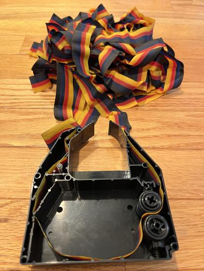 An open empty ImageWriter II color cartridge with the color ribbon in a pile above it with a single loop threaded in. 