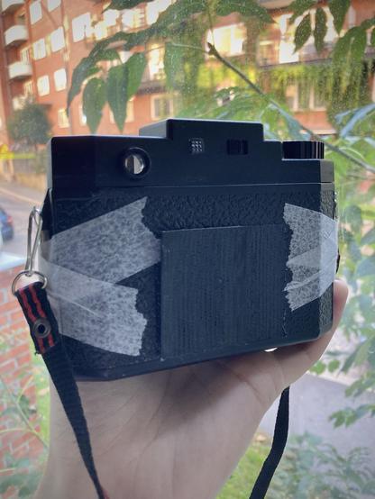 A taped up Holga camera held by me in front of a window. Blurry city background.