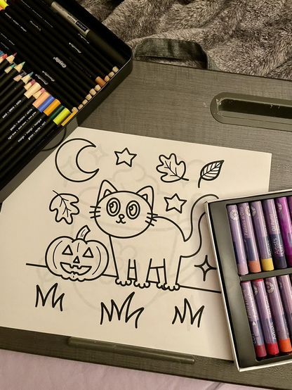 Black and white coloring pages of a cat, carved pumpkin, autumn leaves, stars, and a crescent moon. Pastels in a box bottom right, a tin of colored pencils top left corner. Coloring page by Rana Awadallah 