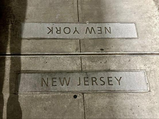 Photo of a sidewalk joint with markings on this side saying "New Jersey" and on the other side "New York".