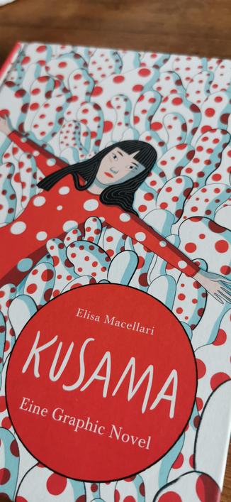 Ansicht vom Cover der Graphic Novel "Kusama"
Inmitten einer Landschaft von wobbeligen Figuren in weiß mit roten Punkten liegt eine japanische Person in einem roten Einteiler, der weiße Punkte hat