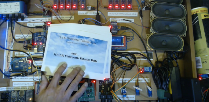 Photo of the Lo-Fi Orchestra, which is a whole pile of various Arduinos and speakers hooked up via MIDI, with a card over the top with a background of white clouds on a blue sky and the words "and MT32-Pi Fluidsynth Tubular Bells".