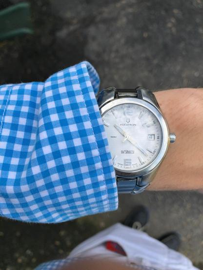 Close-up of a wrist wearing a silver Accutron watch. The wrist is covered by a blue and white gingham shirt, with part of a plaid shirt or pants visible at the bottom of the image. 