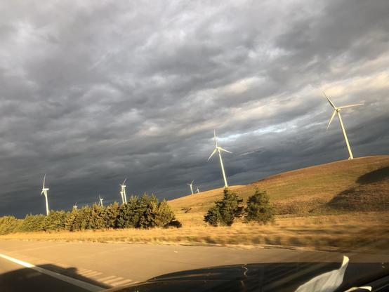A landscape featuring several wind turbines on rolling hills, with a cloudy sky above. The foreground includes grass and trees alongside a highway.