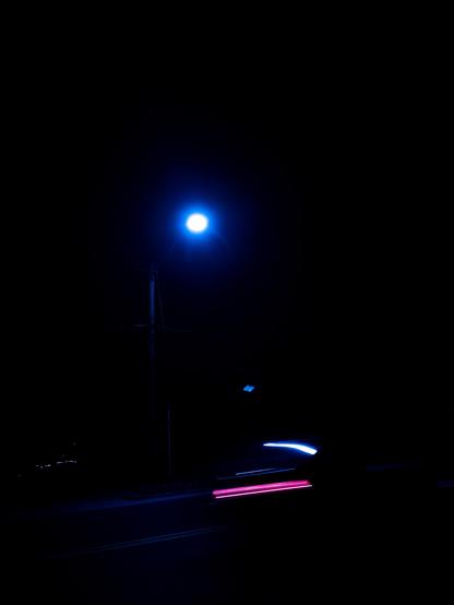A bright blue streetlight illuminates a dark scene, with streaks of light from a passing vehicle in the foreground. The background is largely black, creating a stark contrast.