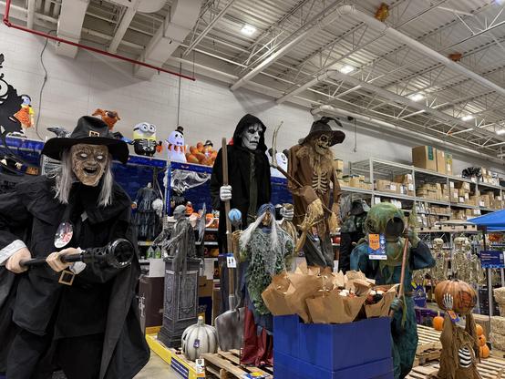 A display of Halloween decorations featuring several life-sized, animated figures, including a witch, a skeletal figure, and various spooky characters. The background includes a range of Halloween-themed items and decorations.