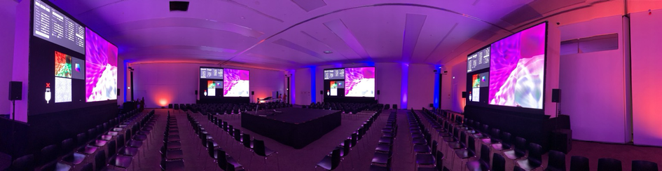 A panoramic photo of a presentation room at the conference center in Tróia, Portugal. There are huge LED screens on all four walls, and the walls themselves are lit with vivid pink, orange and purple light. A stage in the center is surrounded by seats only five rows deep, so everyone in the audience has a great view. 