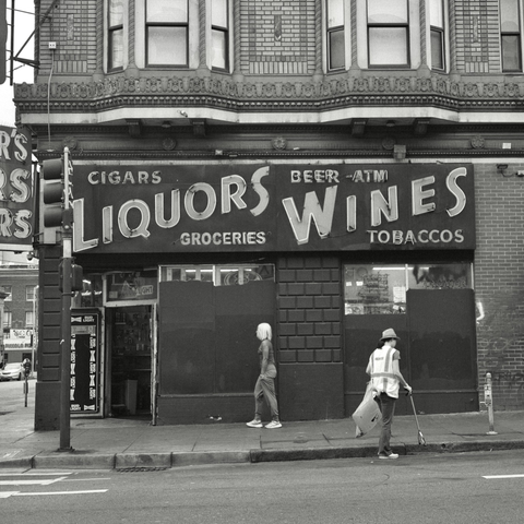 A black and white film photo. Two people walk underneath an old and large grocery store sign in San Francisco, California. Saturday, June 8, 2024.