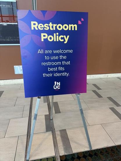 Purple placard with yellow type sitting on an easel in front of a set of restrooms in a convention center. It reads:

Restroom Policy
All are welcome to use the restroom that best fits their identity