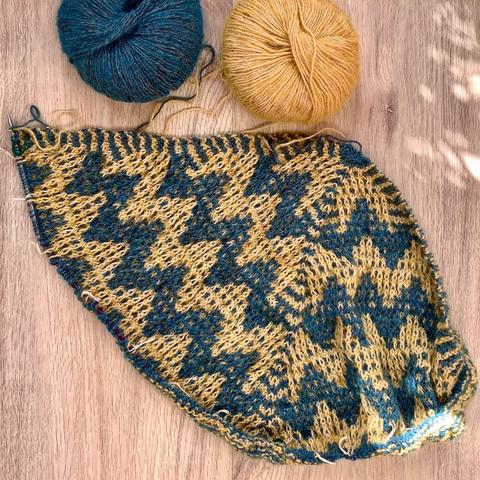 Flat lay view of a triangular shawl with mosaic patterning being knit on a circular needle. It’s shown with a ball of yellow and a ball of petrol colored yarn on a light wooden background.