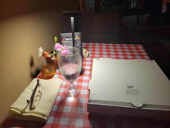 Nighttime photo of a rectangular outdoor cafe table covered in red and white checkered vinyl tablecloth, with the following objects on top: folded off-white cloth napkin with a collapsed pair of glasses resting on top; a small unlit orange-colored glass tea-light holder; a small vase with several cut flowers; a small black table tent menu; a small black illuminated 12" high downward-casting LED lamp with circular base & top connected by a thin stem; a partially-full water glass; a white to-go pizza box. 

The light from the lamp only illuminates the table, leaving the rest of the outdoor patio in relative darkness (though it is partially illuminated from the restaurant and their own table lamps)