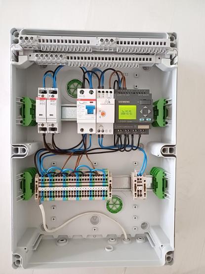 An open damp-proof distribution box with various electrical components and wiring.

In the top row, two ABB contactors are on the left, followed by a 2-pole RCD and a controller for a light sensor. On the right is a Siemens LOGO! controller with a green-lit display showing 'Su 00:00 2008-01-01'.

The bottom row contains Weidmüller terminal blocks in grey, blue, and green-yellow, connected by black, blue and brown wires.