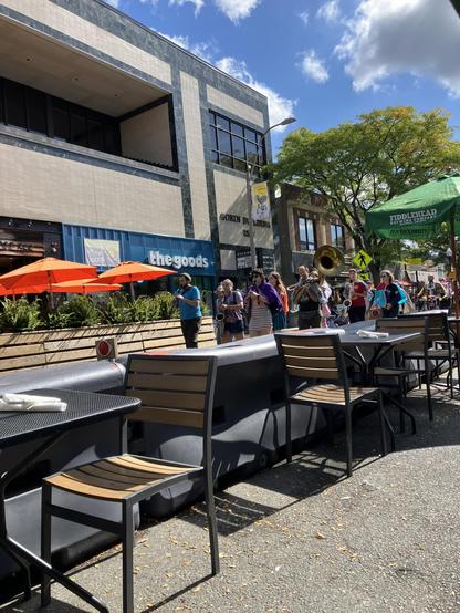 A small marching band walks down a pedestrianized street past a mostly empty patio. 