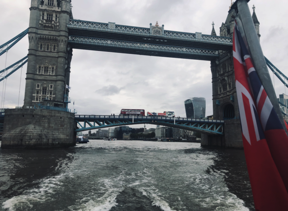 Photo shot from a boat that's just passed through Tower Bridge, leaving a churned-up wake. In the distance, a big red London bus crosses Tower Bridge. 