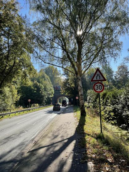 Straße mit Fußweg am Ortsausgang Kessin. Im Hintergrund die Brücke der Bahnstrecke Rostock - Tessin, über die von rechts ein roter Desiro Classic von DB Regio kommt (schwer erkennbar).