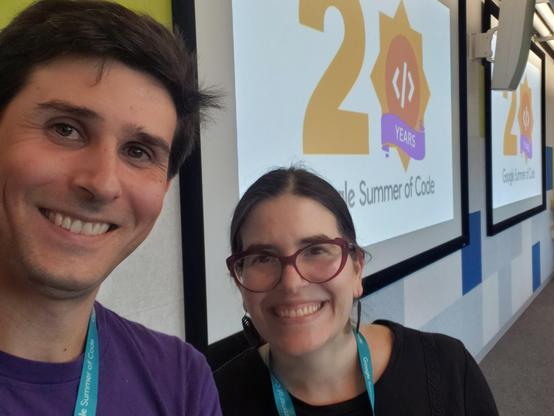 Devin Ulibarri and Karen Sandler smiling in front of a projector screen that says "20 Google Summer of Code".