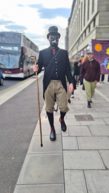 Man with beard dressed in light tweed plus fours, a black top hat &plague doctors mask carrying a bird headed wooden staff, he has black and red boots, red tartan flashes on his long black socks, a black coat with tails, silver buttoned waistcoat and a tartan tie that matches the flashes, street scene behind him including double decker bus