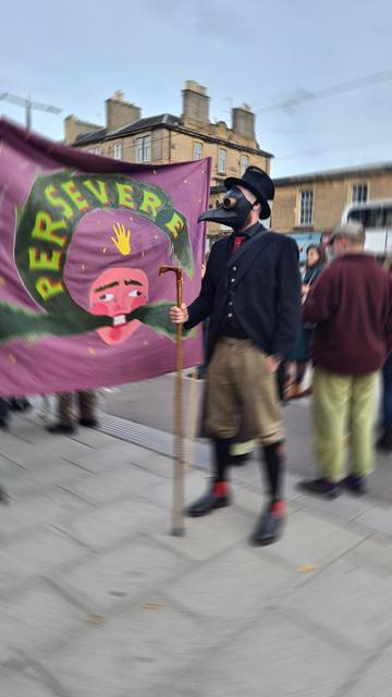 Man with beard dressed in light tweed plus fours, a black top hat &plague doctors mask carrying a bird headed wooden staff, he has black and red boots, red tartan flashes on his long black socks, a black coat with tails, silver buttoned waistcoat and a tartan tie that matches the flashes, purple banner with Folklore flourishes saying Persevere behind him