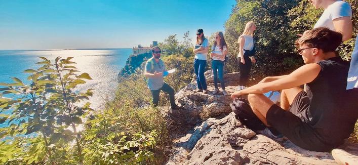The excursion group at the cliffs of Duino. Prof. Tobias Hank is standing close to the edge and is talking to the group. Below, the blue Mediterranean Sea can be seen. There's green shrubbery around the group.
